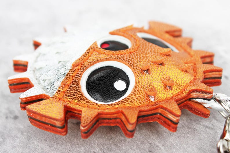 May include: A pufferfish-shaped keychain crafted from layered orange and silver textured material. The design features large, circular black eyes with white pupils and a jagged, spiky perimeter. A metal ring is attached to the side of the fish.