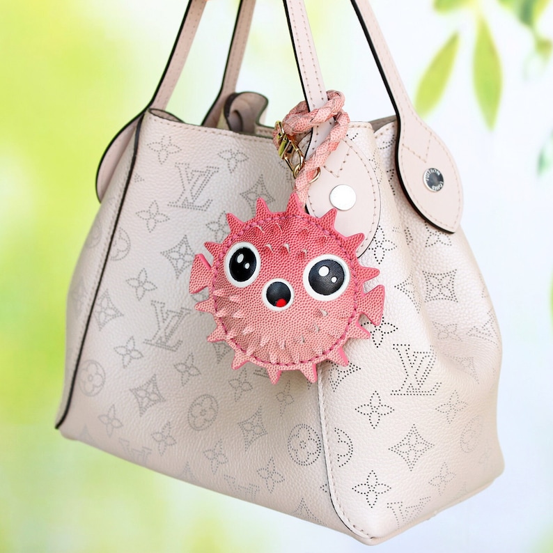 May include: A beige handbag with a repeating monogram pattern and a pink pufferfish-shaped charm attached to the handle. The charm features large black and white eyes, a small open mouth, and textured spikes around its circular body.