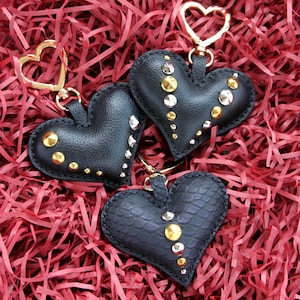 Puede incluir: Tres llaveros negros en forma de corazón con tachuelas doradas y plateadas. Los llaveros tienen cierres dorados en forma de corazón. Los llaveros se exhiben sobre un lecho de papel rojo triturado.