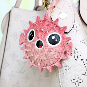May include: A pink pufferfish bag charm with a textured surface, large black and white eyes, and a small mouth with a red interior. The charm features pointed spikes around its circular body and is attached to a light pink handbag with a monogram pattern.