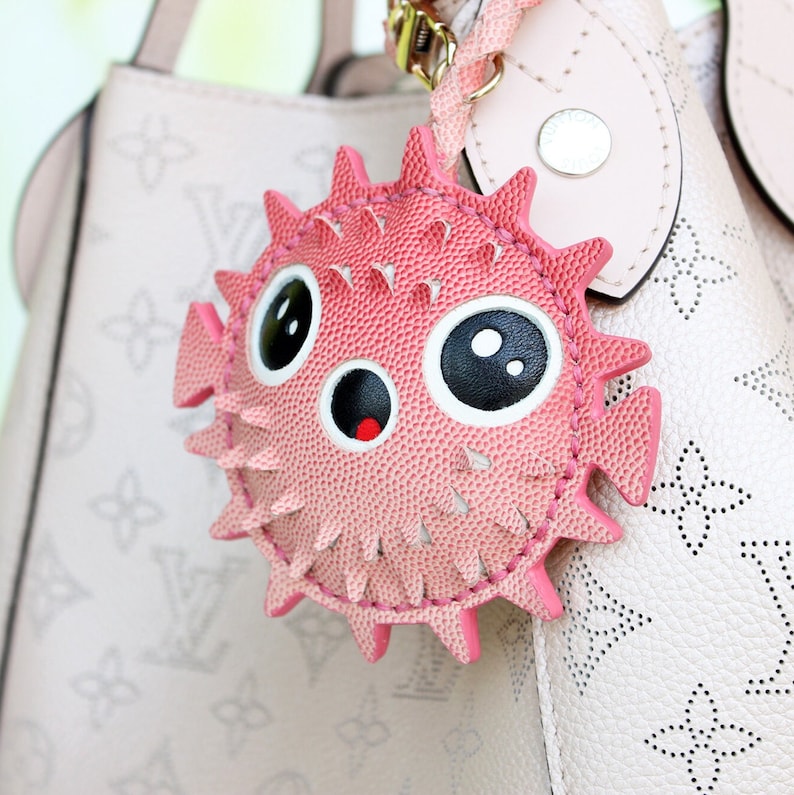 May include: A pink pufferfish bag charm with a textured surface, large black and white eyes, and a small mouth with a red interior. The charm features pointed spikes around its circular body and is attached to a light pink handbag with a monogram pattern.