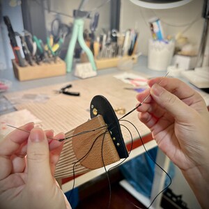 May include: A leather craft project featuring a tan and black leather piece being hand-stitched with a needle and black thread. The leather is secured on a wooden stitching pony, showing visible metal rivets and precise pre-punched holes for sewing.