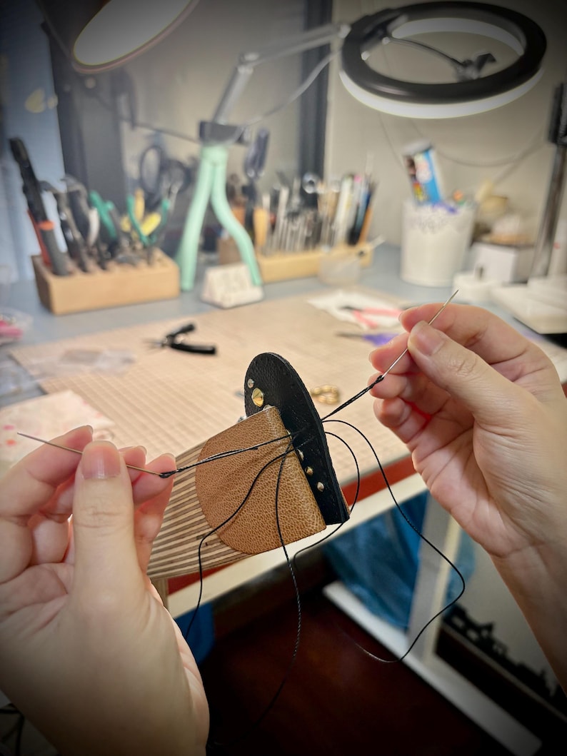 May include: A leather craft project featuring a tan and black leather piece being hand-stitched with a needle and black thread. The leather is secured on a wooden stitching pony, showing visible metal rivets and precise pre-punched holes for sewing.