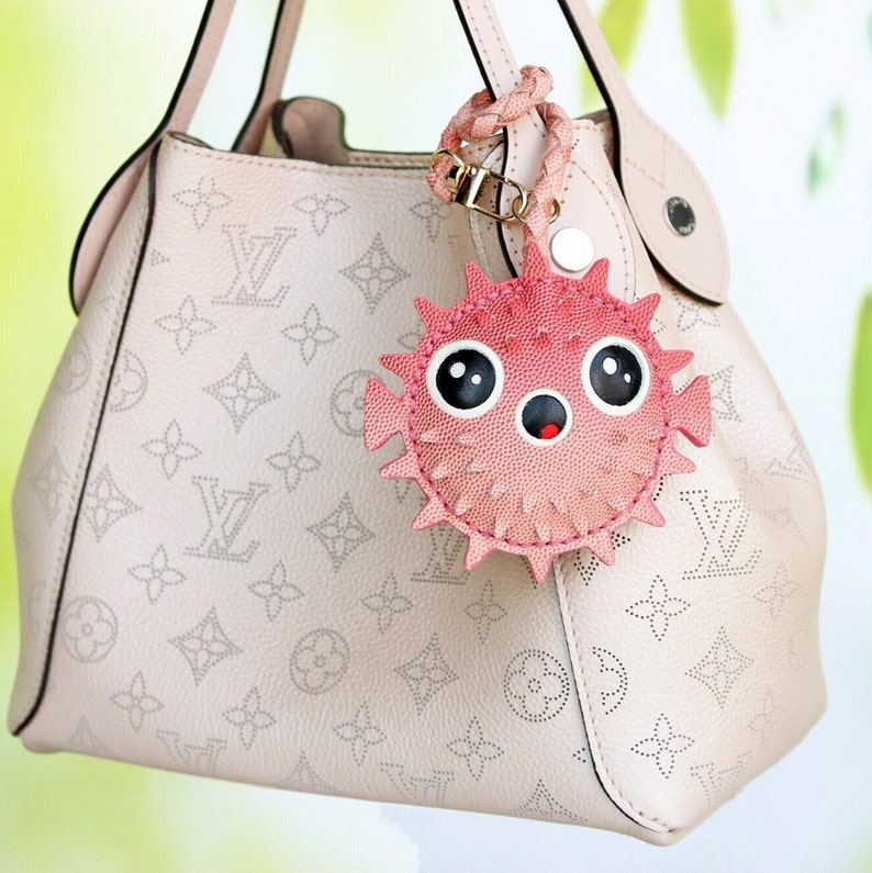 May include: A light beige leather handbag with a perforated monogram pattern and a pink pufferfish-shaped bag charm. The charm features a braided strap, large black and white eyes, and a small circular mouth with a red interior detail.