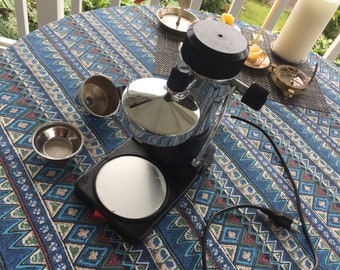 May include: A silver and black electric coffee maker with a chrome drip tray and a black cord. The coffee maker is on a blue and white patterned tablecloth.