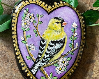 May include: Hand-painted rock art featuring a yellow bird with black and white markings perched on a branch with white flowers. The bird is set within a purple heart-shaped design, framed by a gold-dotted border on a black background.