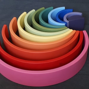 Peut inclure: Un ensemble d'arches en bois arc-en-ciel, disposées en demi-cercle, la plus grande en bas et la plus petite en haut. Les arches sont peintes dans des tons de rose, rouge, orange, jaune, vert, bleu et violet.