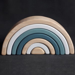 May include: A wooden rainbow stacking toy with seven arches in shades of blue and white. The arches are arranged in a semi-circle, with the largest arch at the bottom and the smallest arch at the top.