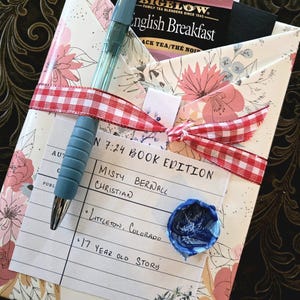 Può includere: Un regalo avvolto in carta floreale, con un nastro a scacchi rosso e bianco e una penna blu. Il regalo include una bustina di tè Bigelow English Breakfast e una nota scritta a mano con il testo "N 7:24 BOOK EDITION".