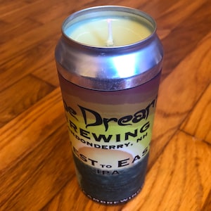 Może przedstawiać: Świeca w srebrnym puszce z żółtym woskiem na górze i knotem. Puszka ma etykietę z tekstem "The Dream Brewing Londonderry, NH West to East IPA".