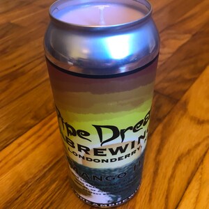 Può includere: Una candela in una lattina d'argento con la scritta "Pipe Dream Brewing Londonderry Mango IPA". La candela ha una cera rosa chiaro e uno stoppino bianco. La lattina presenta un design sfumato con colori giallo, arancione e marrone.