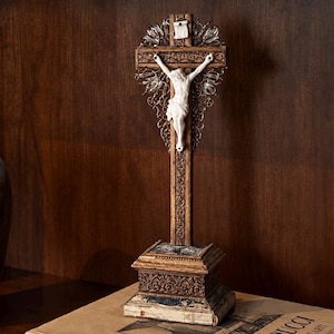 Pode incluir: Um crucifixo de pé com uma figura branca de Jesus montada numa cruz de madeira escura e ornamentada com entalhes intrincados. A cruz é acentuada com elementos de filigrana metálica e repousa sobre uma base retangular escalonada com detalhes.