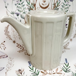 May include: A cream-colored ceramic teapot with a lid and a handle. The teapot has a long spout and a ribbed design on the body. The background features a floral pattern.