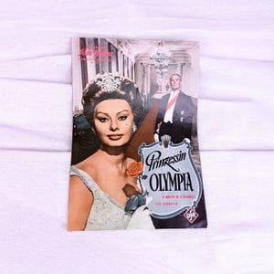Könnte beinhalten: Vintage Filmplakat für "Prinzessin Olympia" mit einer glamourösen Frau mit Tiara und einem Mann in formeller Kleidung. Das Plakat hat einen Vintage-Look mit deutschem Text und einer Rose.