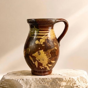 Può includere: Una brocca in ceramica marrone scuro con corpo arrotondato, un manico curvo e un bordo svasato. La superficie è decorata con irregolari schizzi di vernice color giallo chiaro e fasce orizzontali attorno alla zona del collo.