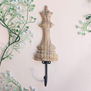 Könnte beinhalten: Ein Wandhaken in Form einer Holz-Schachkönigin mit einem schwarzen Metallhaken. Die Königin ist hellbraun mit dunkelbrauner Maserung. Der Haken ist an einer hellbeigen Wand angebracht, mit weißen und grünen floralen Akzenten.
