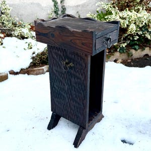 Könnte beinhalten: Ein dunkelbrauner Holzständer mit einer Schublade und einem geschnitzten Rautenmuster. Der Ständer hat eine rechteckige Oberseite mit erhabenem Rand und eine kleine Schublade mit einem Metallringgriff. Der Ständer wird von vier abgewinkelten Beinen getragen.