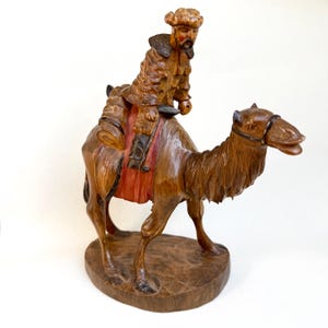 Op de afbeelding: Handgesneden houten beeldje dat een man op een kameel afbeeldt. De man draagt een gedetailleerde jas en hoed. De kameel is bruin met gedetailleerde gravures. Het beeldje is gemonteerd op een houten voet. Het thema is religieus of historisch.