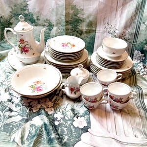Könnte beinhalten: Eine Sammlung von Vintage-Porzellan-Geschirr, darunter eine Kaffeekanne, Teller, Schalen, Tassen und eine Zuckerdose. Die weißen Stücke weisen florale Designs in Rosa, Rot und Gelb mit Goldverzierungen auf. Das Set ist auf einer floral gemusterten Tischdecke angeordnet.