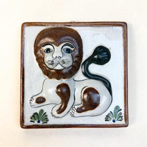 Puede incluir: Azulejo cerámico cuadrado con diseño de león. El león tiene melena, cola y manchas marrones, con cuerpo y cara blancos. El azulejo tiene un borde marrón y detalles decorativos verdes y azules. El león está sentado.