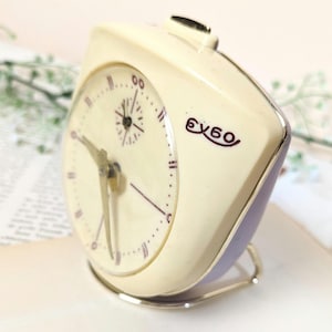 Op de afbeelding: Een witte vintage wekker met een paarse basis en een zilveren standaard. De wijzerplaat heeft zwarte Romeinse cijfers als uurmarkeringen en een kleine secondewijzer. De klok is gelabeld "eybo" in rood.