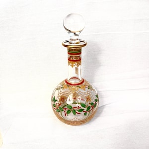 Könnte beinhalten: Eine klare Glasdekanter mit rundem Boden und langem Hals, gekrönt von einem klaren Glasverschluss. Der Dekanter ist mit goldenen, roten und grünen Blumen- und Blättermustern verziert. Der Hals hat rote und grüne Bänder.