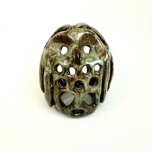 Può includere: Una scultura in ceramica dal design unico. Il pezzo presenta una forma arrotondata con molteplici aperture circolari. La combinazione di colori include sfumature di verde e marrone, con una finitura lucida. La scultura ha una superficie strutturata ed è probabilmente fatta a mano.