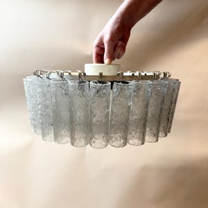 Puede incluir: Una lámpara de araña vintage con un diseño circular. Cuenta con una serie de cilindros de vidrio transparente texturizado suspendidos de un marco de metal. La lámpara tiene un componente central de color crema y está sostenida por una mano.