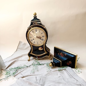 Puede incluir: Un reloj negro antiguo con detalles dorados y una esfera crema. El reloj presenta números romanos y un péndulo. También hay una caja decorativa negra y dorada a juego y un pequeño soporte con un pomo dorado.