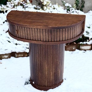 Op de afbeelding: Een donkerbruine houten tafel met een halfronde bovenkant en een cilindrische voet. De bovenkant van de tafel heeft een gebogen voorkant met verticale houten latten. De voet is ook gemaakt van verticale houten latten. De tafel staat buiten in een besneeuwde omgeving.