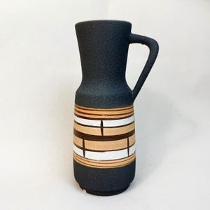 Può includere: Brocca in ceramica nera con manico. La parte inferiore presenta fasce orizzontali bianche, beige e marroni. La brocca ha una superficie strutturata e un design vintage classico.