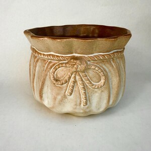 Può includere: Una fioriera in ceramica beige e marrone. Ha la forma di un sacchetto legato con un fiocco di corda. Il bordo e l'interno sono marrone scuro, mentre l'esterno è beige chiaro con dettagli di corda.