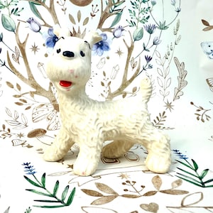 Könnte beinhalten: Vintage weiße Terrier-Hundefigur mit rotem Mund und schwarzen Augen. Der Hund hat ein strukturiertes Fell und steht auf allen Vieren. Der Hintergrund zeigt ein Blumen- und Geweihmuster.