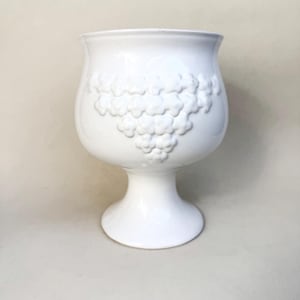 Può includere: Un vaso in ceramica bianca con base a piedistallo e parte superiore ampia e arrotondata. Il vaso presenta un motivo floreale in rilievo sul lato. La forma generale ricorda un calice o un calice, adatto per esporre fiori o come elemento decorativo.