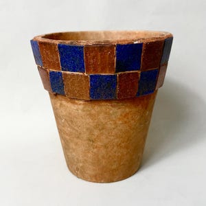 Puede incluir: Una maceta de terracota con una banda de mosaico. La banda presenta un patrón de tablero de ajedrez de baldosas marrones y azules. La maceta tiene una forma ligeramente acampanada y un acabado liso y sin esmaltar.