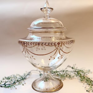 Può includere: Un vaso di vetro trasparente con coperchio, con decorazioni dorate. Il vaso ha una base a piedistallo ed è decorato con un motivo a foglie e nastri dorati attorno al bordo. Il coperchio è sormontato da un pinnacolo appuntito.