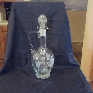 May include: A clear glass decanter with a floral design etched into the glass. The decanter has a stopper with a bulbous top.
