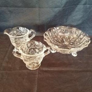 May include: A set of three clear glass serving dishes with an etched floral design. The set includes a large bowl with three feet and two smaller creamer-style dishes.