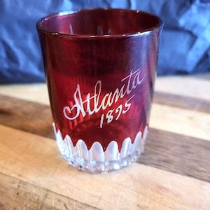 Op de afbeelding: Een rood glazen shotglas met een wit gegraveerd ontwerp met de tekst "Atlanta 1895".