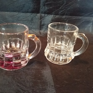 Puede incluir: Dos vasos de chupito de vidrio transparente con asa. El vaso de la izquierda tiene un líquido rojo oscuro en su interior. Ambos vasos tienen un diseño texturizado.