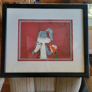 Puede incluir: Un cel de dibujos animados enmarcado de Bugs Bunny, en un esquema de colores azul y gris, con un fondo rojo. Bugs sostiene una zanahoria y parece estar sudando. El cel está enmarcado en un marco negro con un passe-partout blanco.