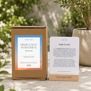 May include: A brown cardboard box labeled Mindfulness Affirmation Motivation with a sunset graphic, next to a white instruction card in a light wood stand. The card text reads How to Use and includes instructions for a Weekly Anchor Practice.