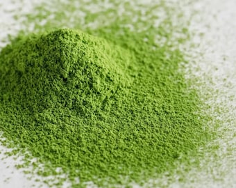 Puede incluir: Primer plano de un montón de polvo de matcha verde brillante. El polvo finamente molido se concentra en un pequeño montículo, con una dispersión del polvo en los bordes. El color verde brillante sugiere frescura y calidad.
