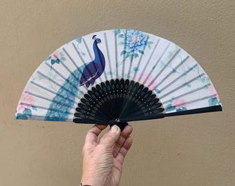 Puede incluir: Abanico de mano decorativo con diseño de pavo real. El abanico tiene un mango de madera negro y una superficie de papel blanco adornada con un pavo real azul, detalles florales rosas y azules y líneas blancas verticales.