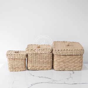 May include: Three woven storage baskets with lids, in various sizes, on a white marble surface. The baskets are made of natural materials and have a light brown colour.