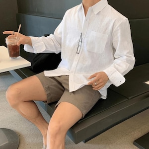 Peut inclure: Une chemise blanche en lin avec une poche poitrine et des manches longues, portée avec un short vert olive. La chemise est déboutonnée en haut, et des lunettes sont suspendues au col. Un verre de café glacé est tenu à la main.