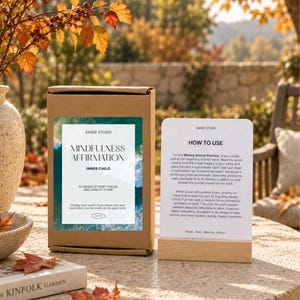 May include: A brown cardboard box labeled Mindfulness Affirmation Inner Child by Sagee Studio, next to a white instruction card in a wooden stand. The card details how to use the weekly anchor practice and daily reality check for quiet focus.