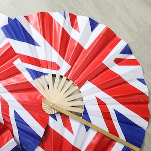 Puede incluir: Varios abanicos abiertos con el diseño de la bandera del Reino Unido, en rojo, blanco y azul. Los abanicos tienen un mango de madera y están parcialmente desplegados, mostrando el patrón de la bandera. Sobre una superficie clara.