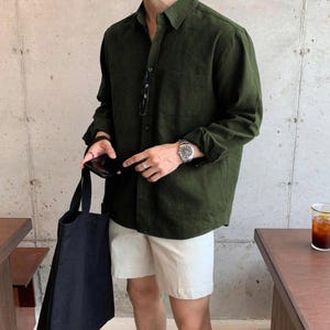 Puede incluir: Camisa verde oscuro de manga larga con botones y bolsillo en el pecho. La camisa se combina con pantalones cortos blancos y un bolso tote negro. Se ve un reloj plateado en la muñeca.