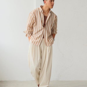 May include: A person wearing a brown and white striped long-sleeved shirt with a white undershirt and beige wide-leg pants.
